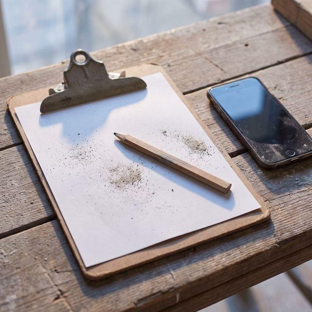 Klemmbrett mit Papier und Bleistift neben einem Smartphone auf einem Holztisch.